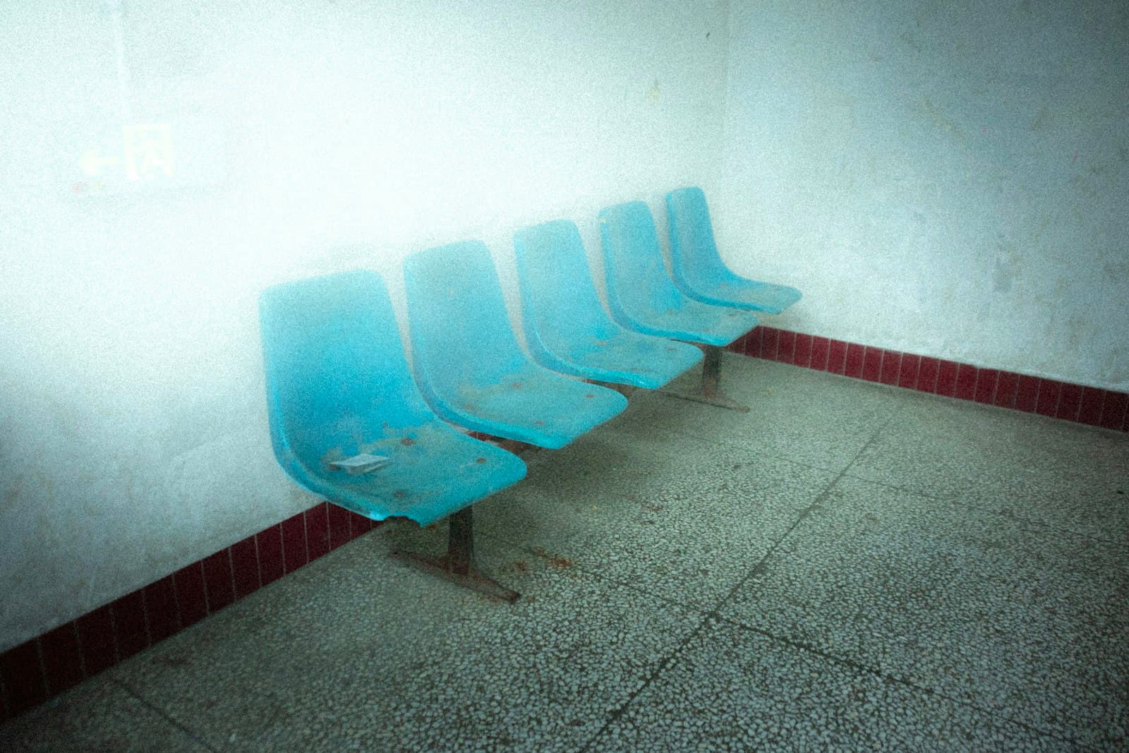 A row of empty blue chairs against the wall of a bare institutional waiting room